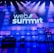 A large stage setup with an illuminated sign reading 'Web Summit' against a backdrop of stacked cube-like structures with purple and blue lighting. Several spotlights shine from above, and there is a seating arrangement that includes chairs and a table.